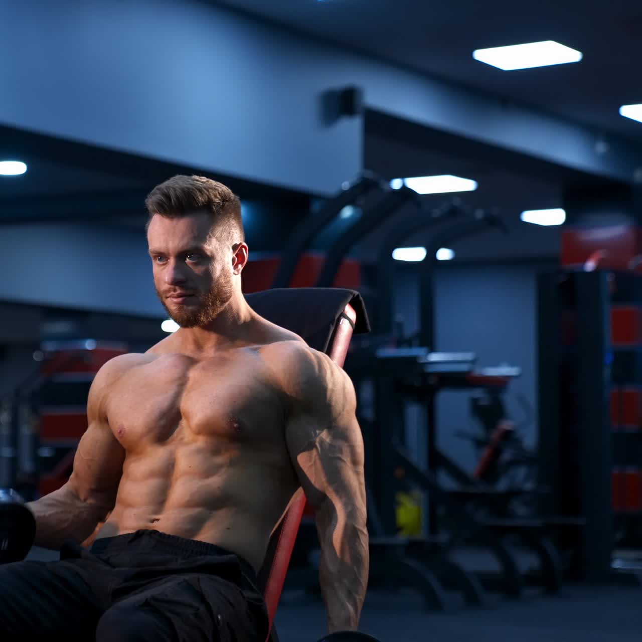 Uomo atletico e affascinante si allena con i manubri su una panca fitness. Sfondo di palestra moderna. Slow motion