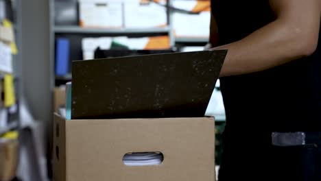 a person wearing a watch and ring is organizing documents in a cardboard box within a file storage room filled with various folders and boxes