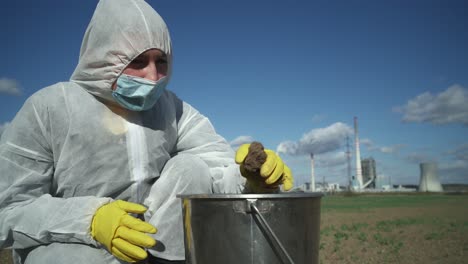 scientist in protective suit checking contamination of field clay, industrial background, ecological disaster concept