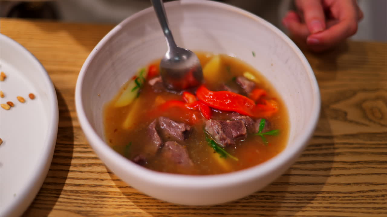 Spoon mixing a broth with chunks of beef, red bell peppers, yellow potatoes, and herbs