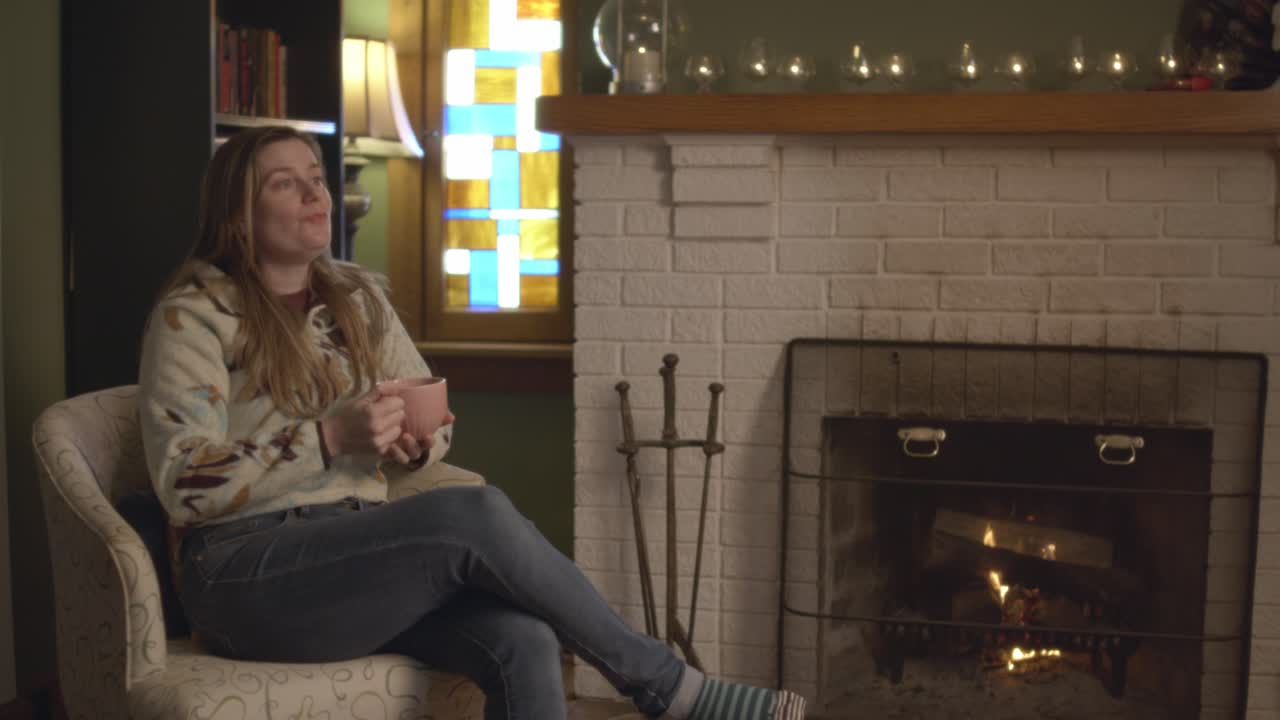A woman in a sweater in a cozy living room sitting, drinking hot tea next to a fireplace. Sipping on tea and relaxing in the warm environment with a crackling fire. Shot in 60 frames for slow motion