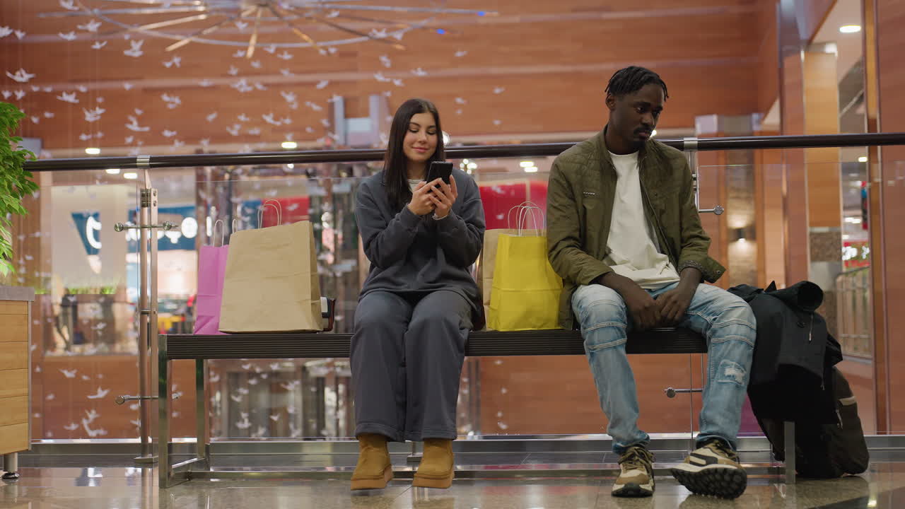 Man looks sad while woman cheerfully operates smartphone beside shopping bags on mall bench, surrounded by modern wooden decor, decorative paper birds, and bright mall lighting