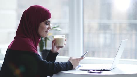 Una-Atractiva-Chica-Musulmana-Con-Hijab-En-La-Cabeza-Está-Bebiendo-Capuchino-Mientras-Busca-Algo-En-Su-Teléfono-Inteligente.-Metraje-En-Interiores