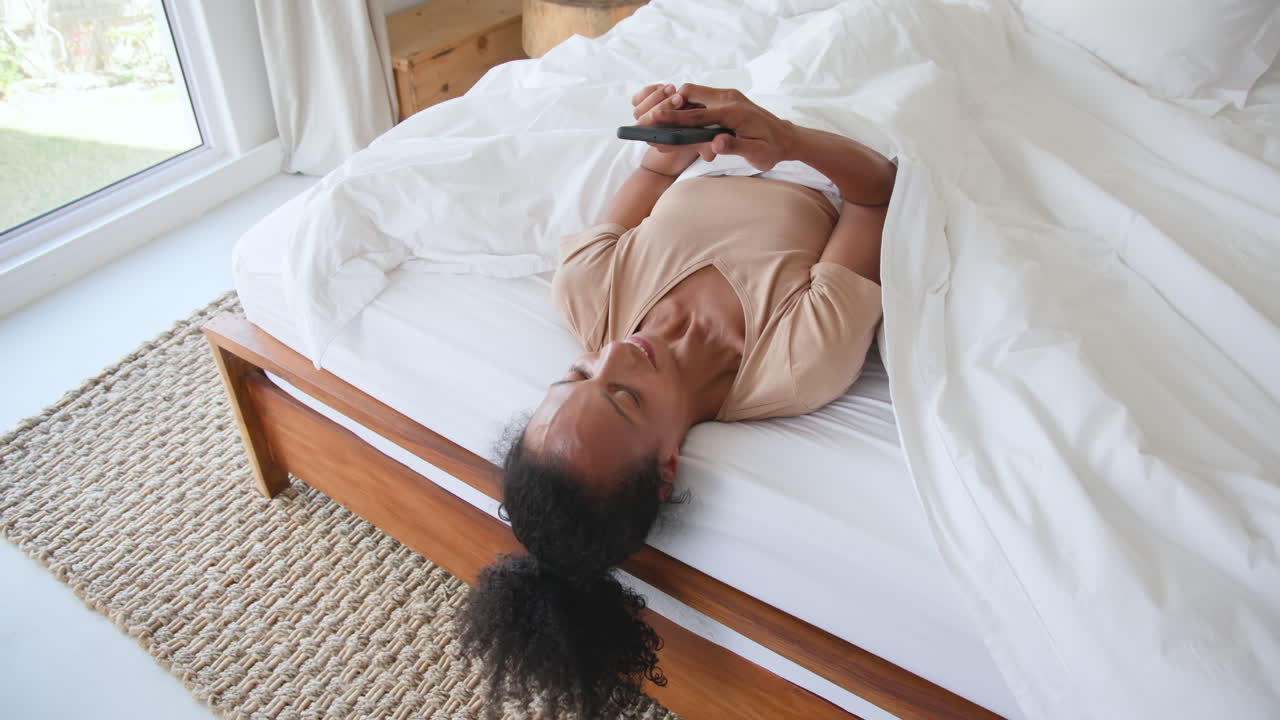 Donna che si rilassa a letto, sorridente mentre guarda un video sullo smartphone, a casa.