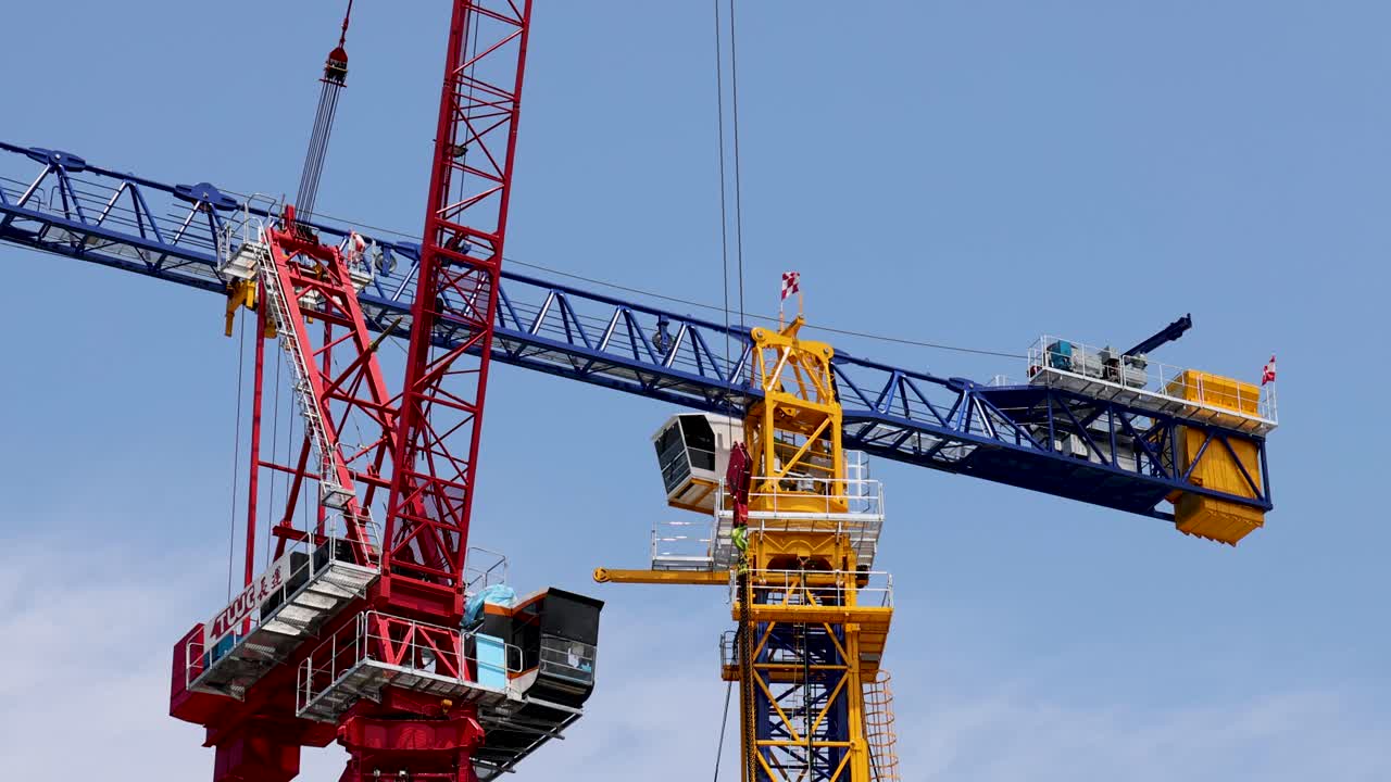 Red tower crane lifts blue boom section vertically, clear daylight, static camera, urban construction environment