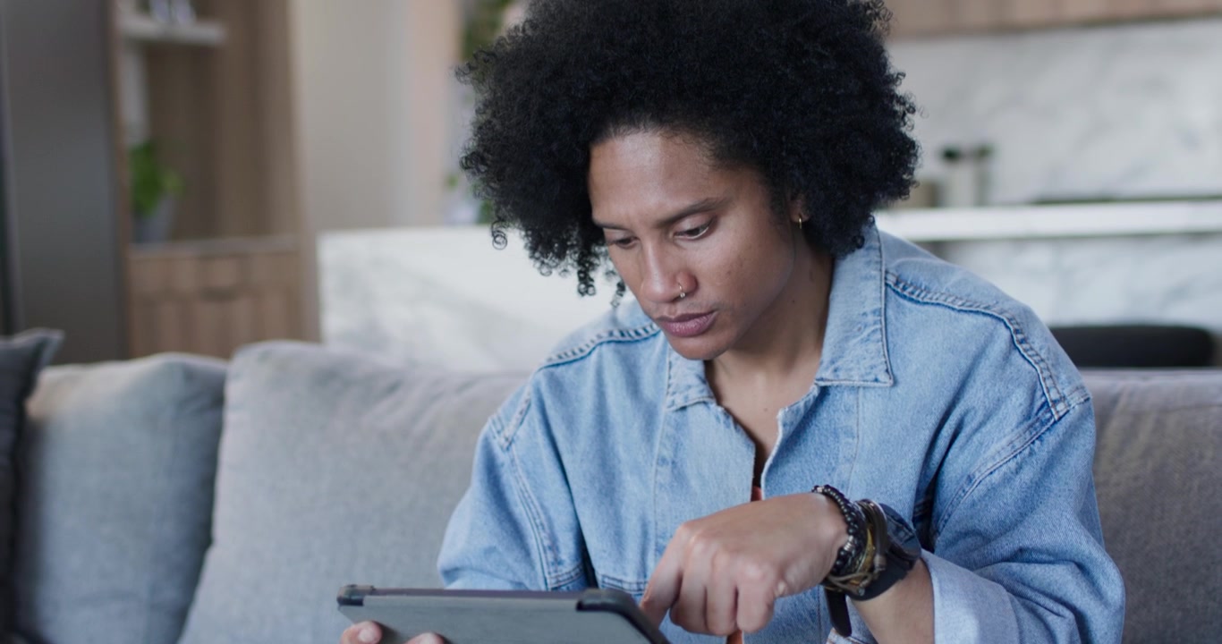 African American man in denim sitting on gray sofa resting chin picking up tablet checking content