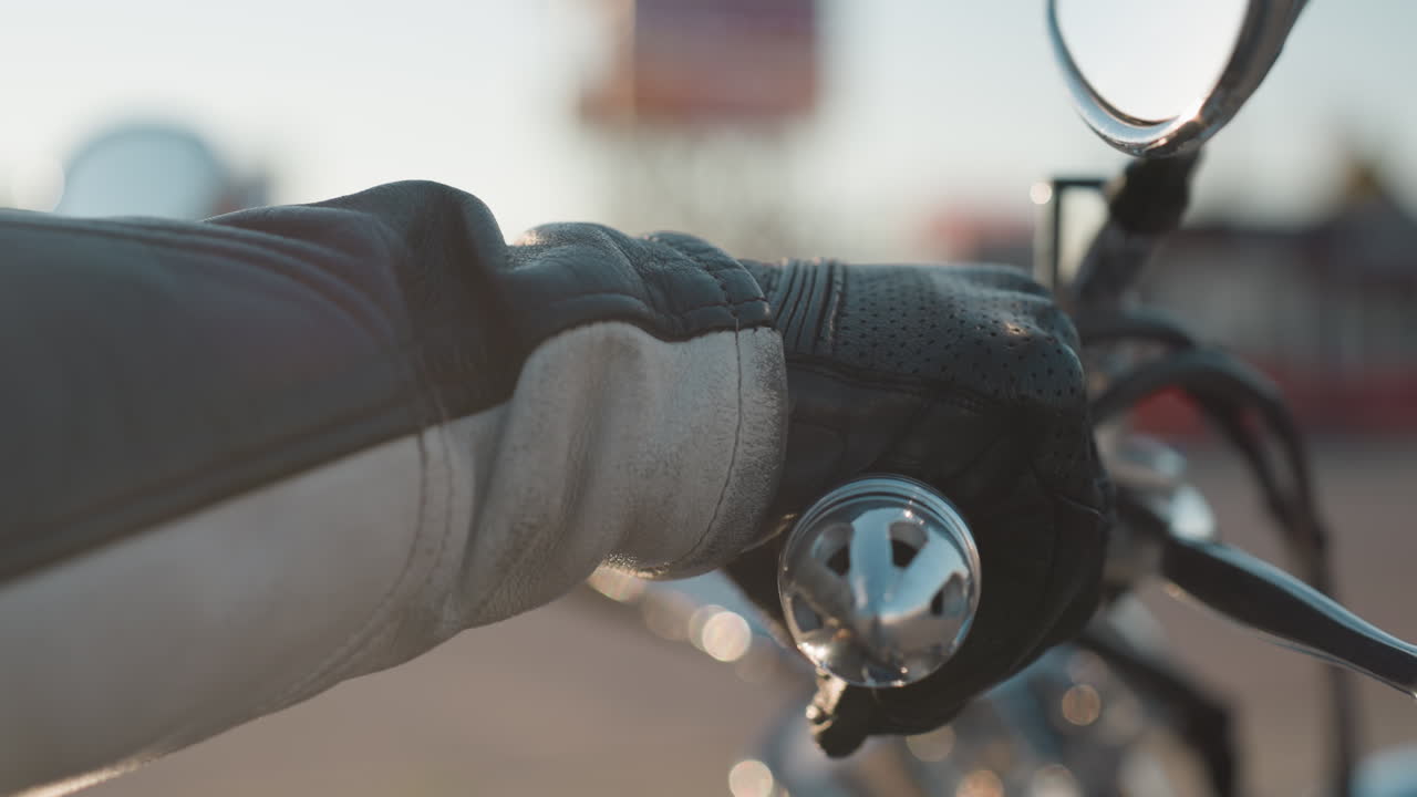 Close up of rider hand on motorcycle handle while throttling, leather glove gripping firmly, chrome handlebar and mirror reflecting blurred city street background