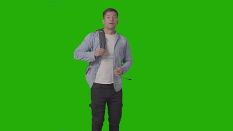 three quarter length shot of smiling male university or college student putting on backpack ready for class against green screen