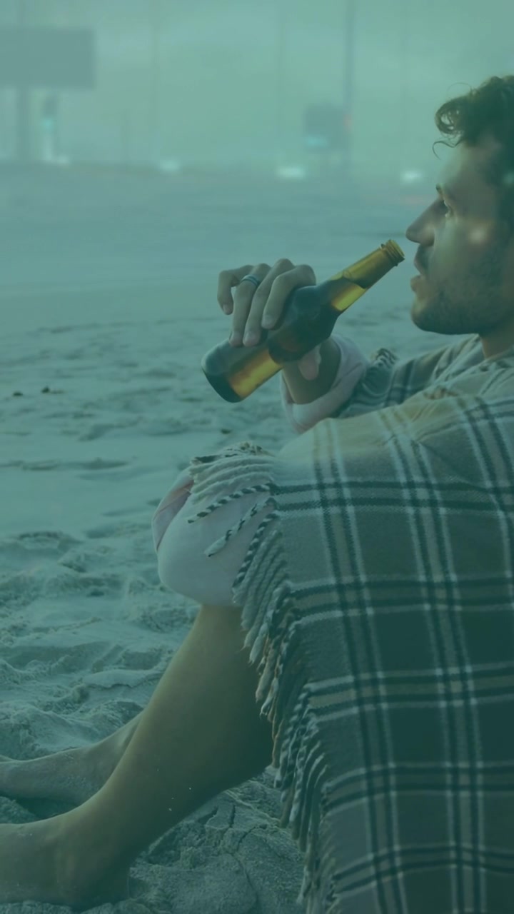 Video vertical: Hombre sentado con camisa de cuadros en la arena, levantando una botella de cerveza, bebiendo a sorbos y contemplando el horizonte en busca de bienestar.