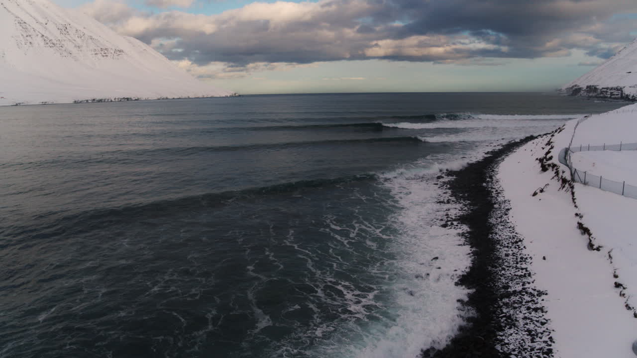 драматическое исландское побережье с заснеженными скалами и разбивающимися океанскими волнами под арктическим небом, дрон спускается в воду на скалах