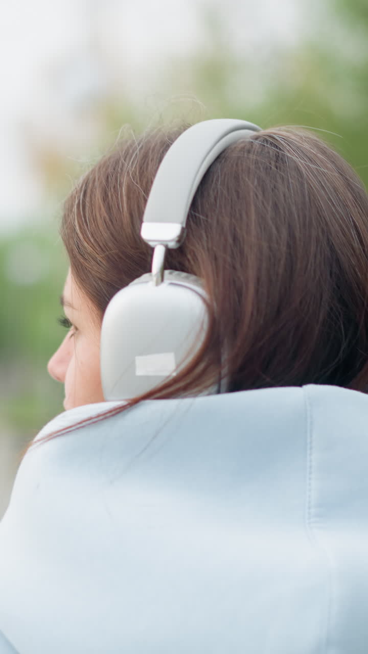 Vista di spalle di una giovane donna con le cuffie che ascolta musica mentre passeggia in un giardino con alberi sfocati sullo sfondo. Ideale per video che mostrano relax, musica e passeggiate nella natura.