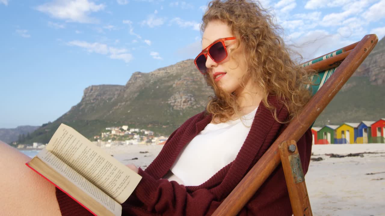Vista lateral de jovem mulher caucasiana com óculos de sol sentada em espreguiçadeira e lendo um livro na praia 4k