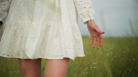 a close-up of a woman's lower body as she strolls through a lush green field, wearing a flowing white dress. her hand gently touches the tall grass