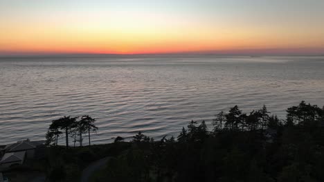 aerial view pulling away from the sunset to reveal a forest and homes overlooking the water