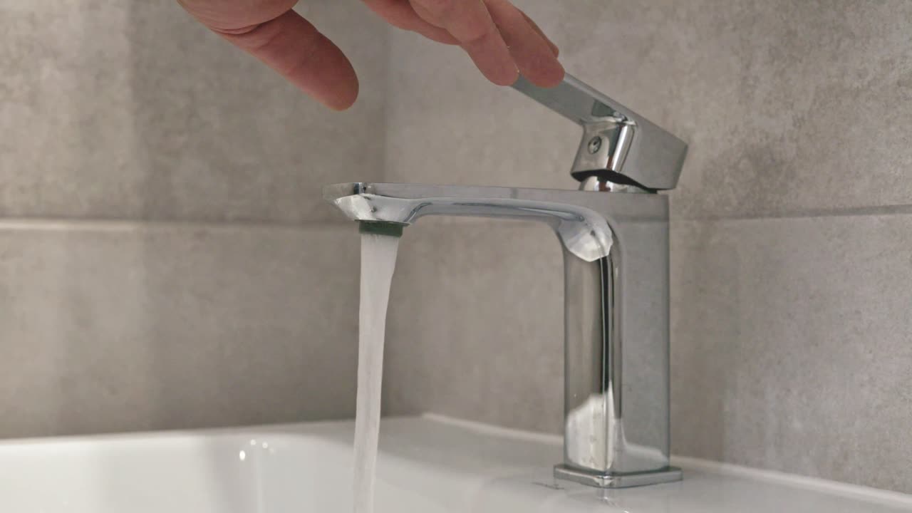 A close-up shot of a modern chrome bathroom faucet with water flowing into a white sink. A hand is adjusting the lever, emphasizing everyday hygiene and water usage in a minimalistic bathroom setting