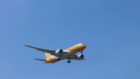 a yellow commercial airplane approaches with landing gear extended against a clear blue sky.