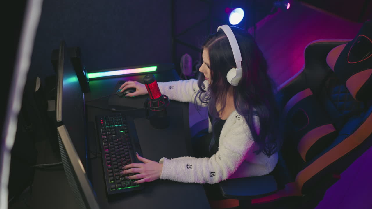 aerial view young girl wearing white headphones designing on computer while seated on gaming chair surrounded by colorful lights, red microphone, and dual monitors in creative tech workspace