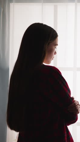 attractive young woman with long flowing hair looks out of french window with light curtain in room zoom in slow motion. waiting for lockdown ending