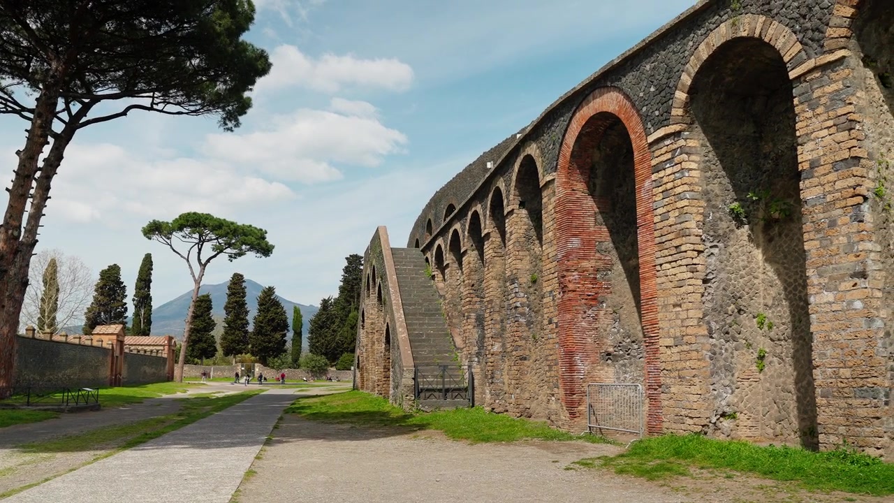 Ydre arkade af Pompeji Amfiteater med murstensbuer, stenmure, trappe, parasolfyrretræer og en solbeskinnet sti gennem det gamle romerske område.
