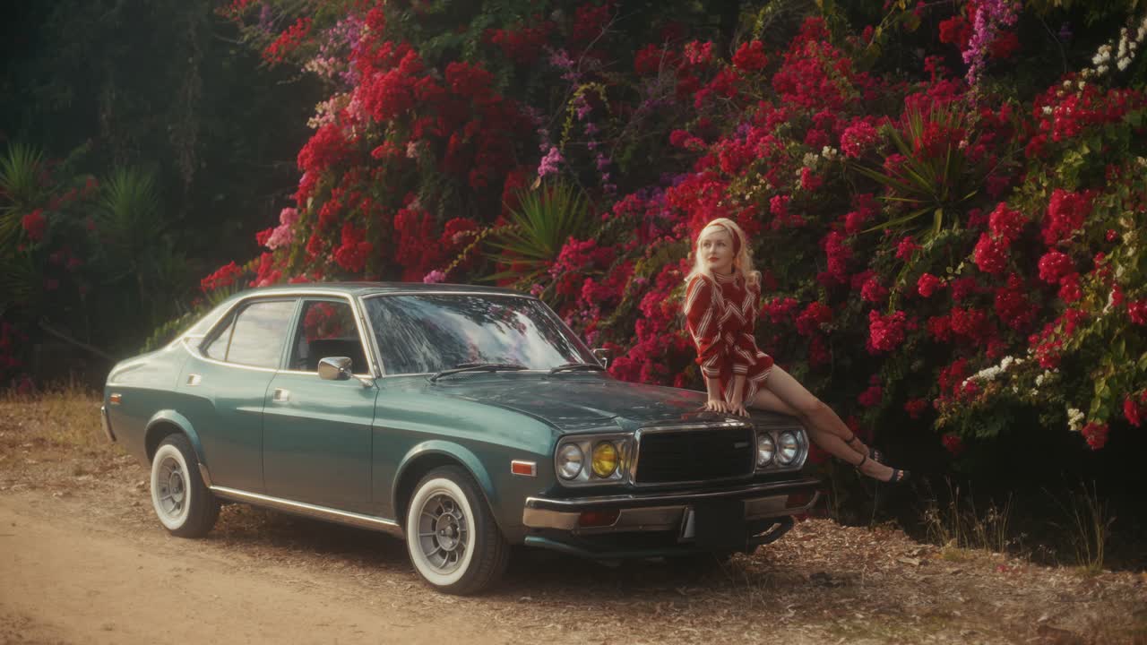 Blonde Frau posiert auf der Motorhaube eines Oldtimers neben roten Blumen