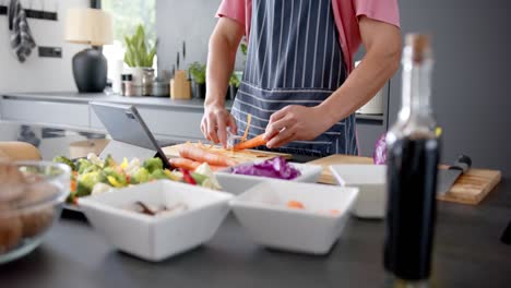 midsection of biracial man in apron preparing vegetables and using tablet in kitchen, in slow motion