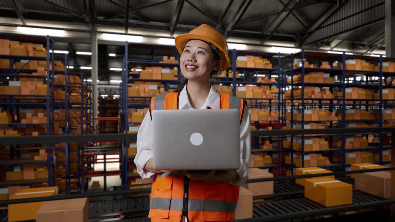 ingeniera asiática con casco de seguridad mirando una computadora portátil y mirando a su alrededor mientras está de pie en el almacén con estantes llenos de productos de entrega