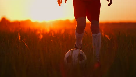 primer plano de seguimiento de los pies de un niño de fútbol en una camiseta roja y pantalones cortos corriendo con la pelota al atardecer en el campo en la hierba. el joven jugador de fútbol sueña con una carrera profesional y se entrena en el campo. en el camino al éxito
