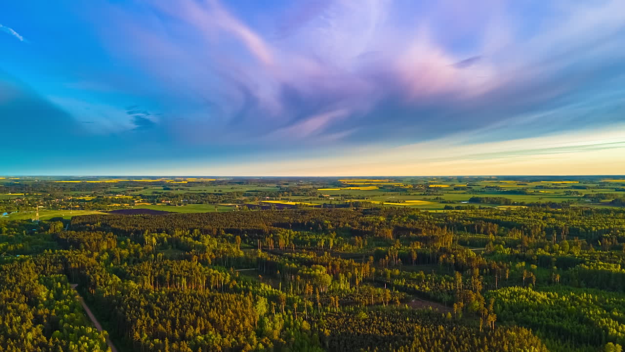 An aerial hyperlapse captures a sweeping forward motion over a diverse Latvian landscape of lush green forests and patchwork fields under a dynamic sky with fast-moving clouds at sunset