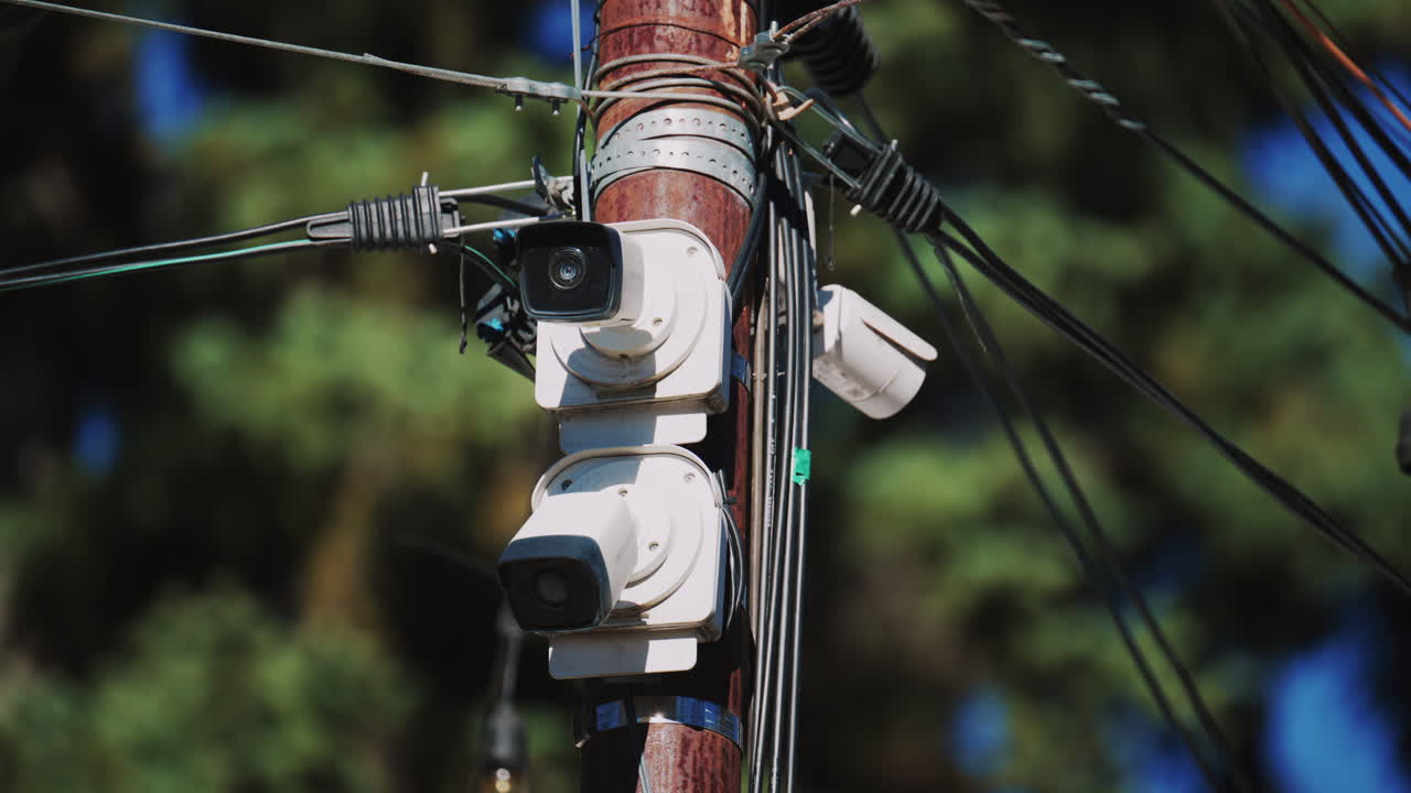 Close up of a utility pole with surveillance cameras, wires, and hanging lights against a blue sky