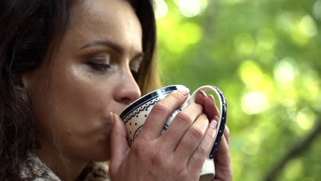 close up fhd shot of a brunette middle-aged woman with her eyes closed savoring a cup of fresh hot tea in the morning with trees in the background