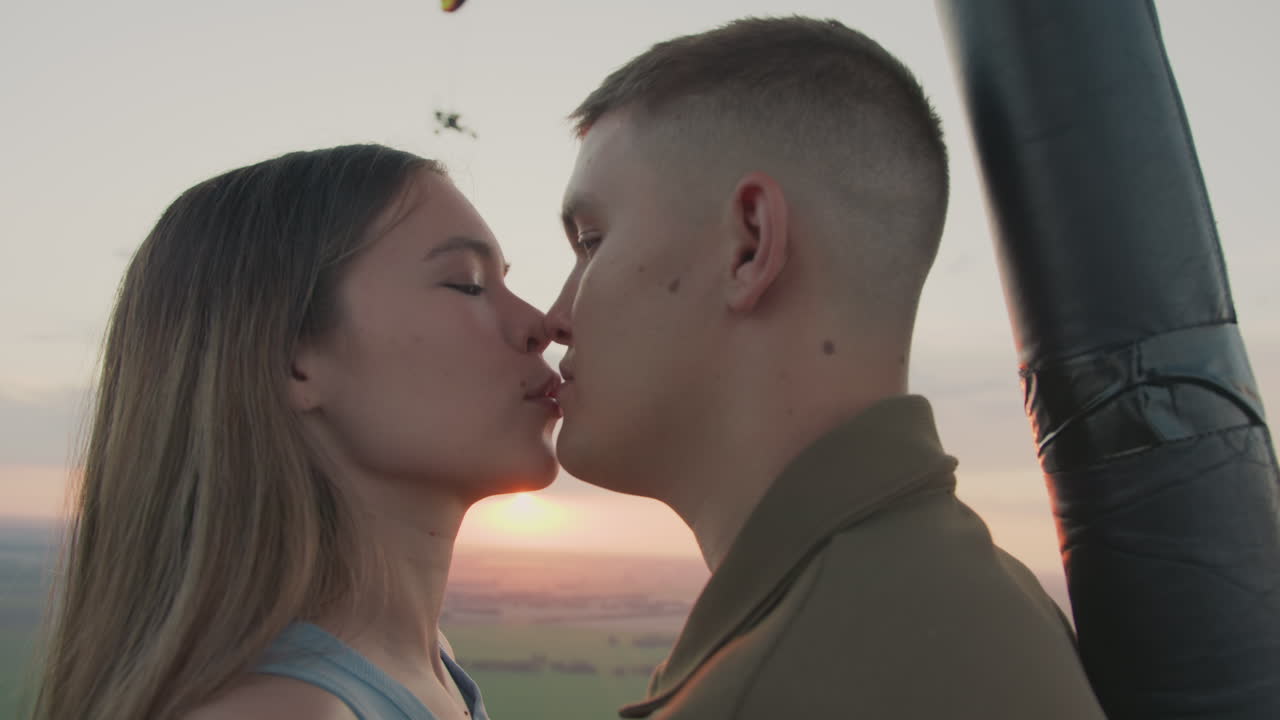 close up lovers in hot air balloon kissing and admiring vibrant sunset sky with distant paraglider drifting over patchwork fields above golden horizon capturing romantic adventure