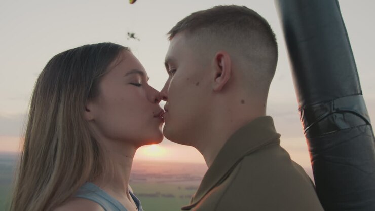 close up lovers in hot air balloon kissing and admiring vibrant sunset sky with distant paraglider drifting over patchwork fields above golden horizon capturing romantic adventure