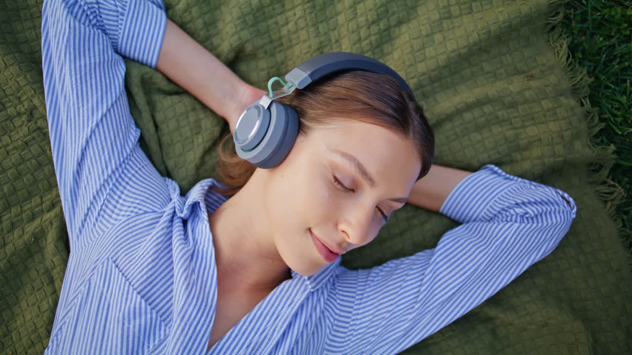 Closed-eyes woman listening music in headphones relaxing picnic blanket closeup