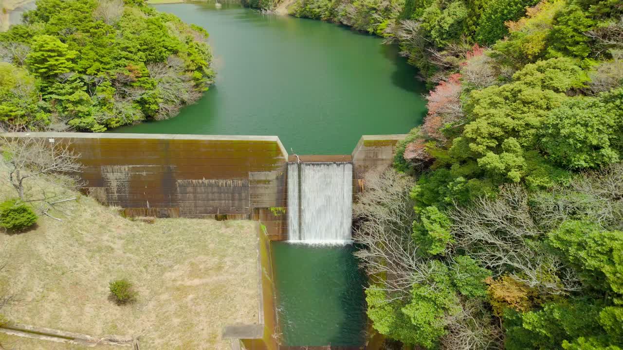 An aerial view pulling back from a concrete dam's spillway to reveal the calm green reservoir and surrounding lush forest. A tranquil scene of water management in a natural setting