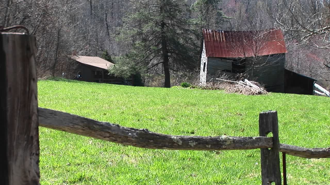 la cámara mira sobre la cerca de riel dividido que enmarca los edificios agrícolas abandonados rodeados de hierba verde brillante