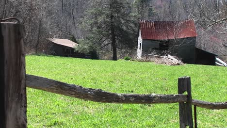 Camera-Peers-Over-Split-Rail-Fence-That-Frames-Abandoned-Farm-Buildings-Surrounded-By-Bright-Green-Grass