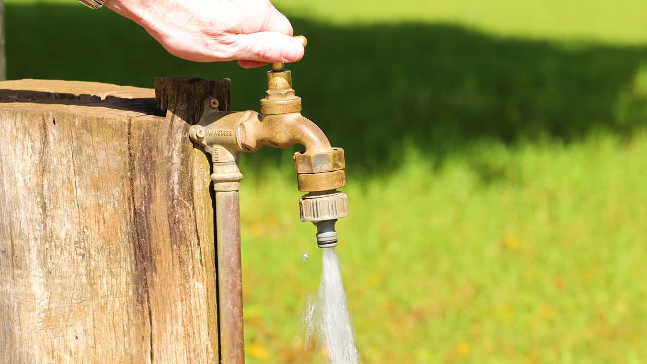 Una mano chiude un rubinetto d'acqua in ottone in una giornata di sole, con l'acqua che scorre su uno sfondo erboso.