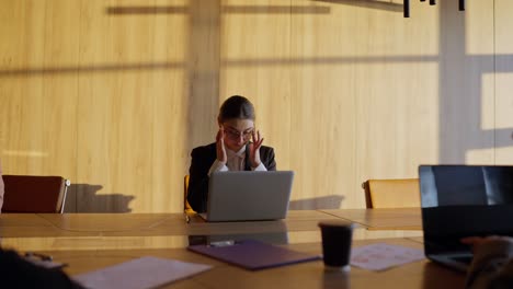 Tired-brunette-businesswoman-in-round-glasses-and-a-black-business-suit-sits-at-a-wooden-table-and-massages-her-temples-during-a-hard-day-at-work-in-a-sunny-modern-office