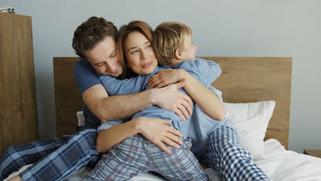 rear of cute little boy hugging his mother and father while they are sitting on the bed in the morning