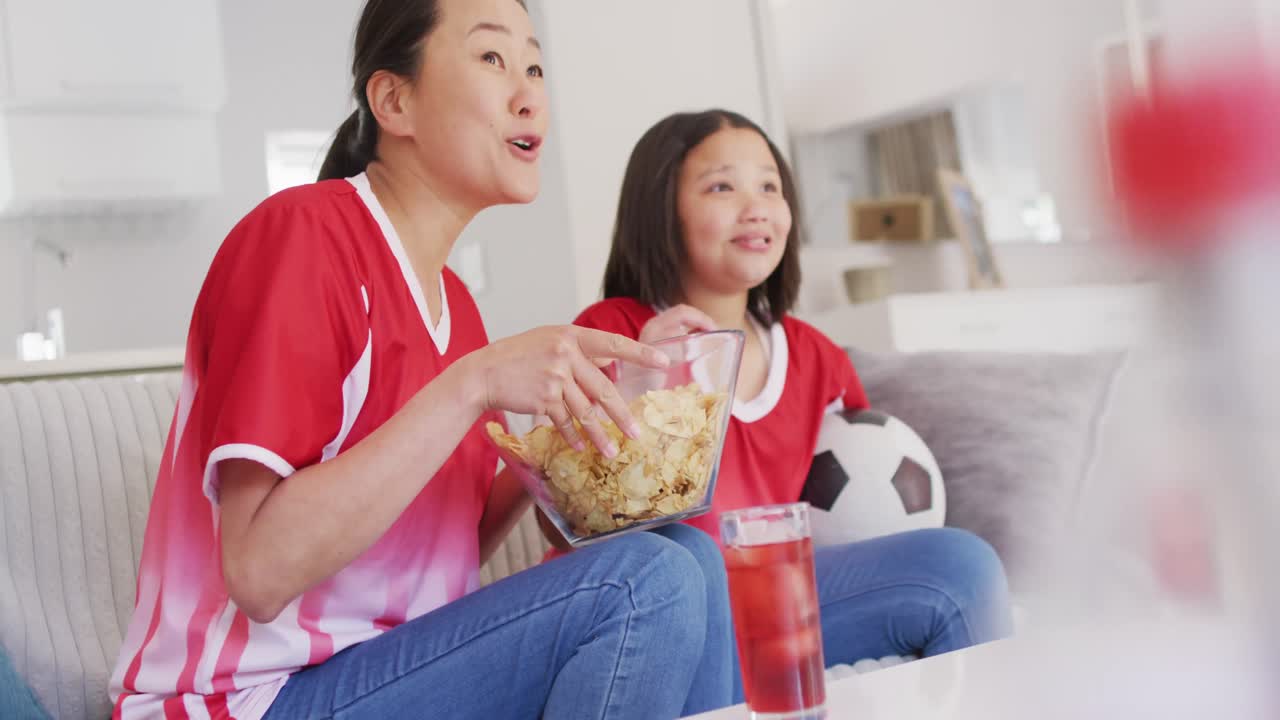 Video di madre e figlia asiatiche felici che mangiano patatine e guardano una partita in TV
