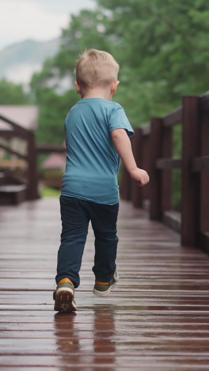 Happy little blond boy runs turning back along wet veranda deck at wooden cottage house after warm spring rain backside view slow motion