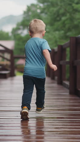 happy little blond boy runs turning back along wet veranda deck at wooden cottage house after warm spring rain backside view slow motion