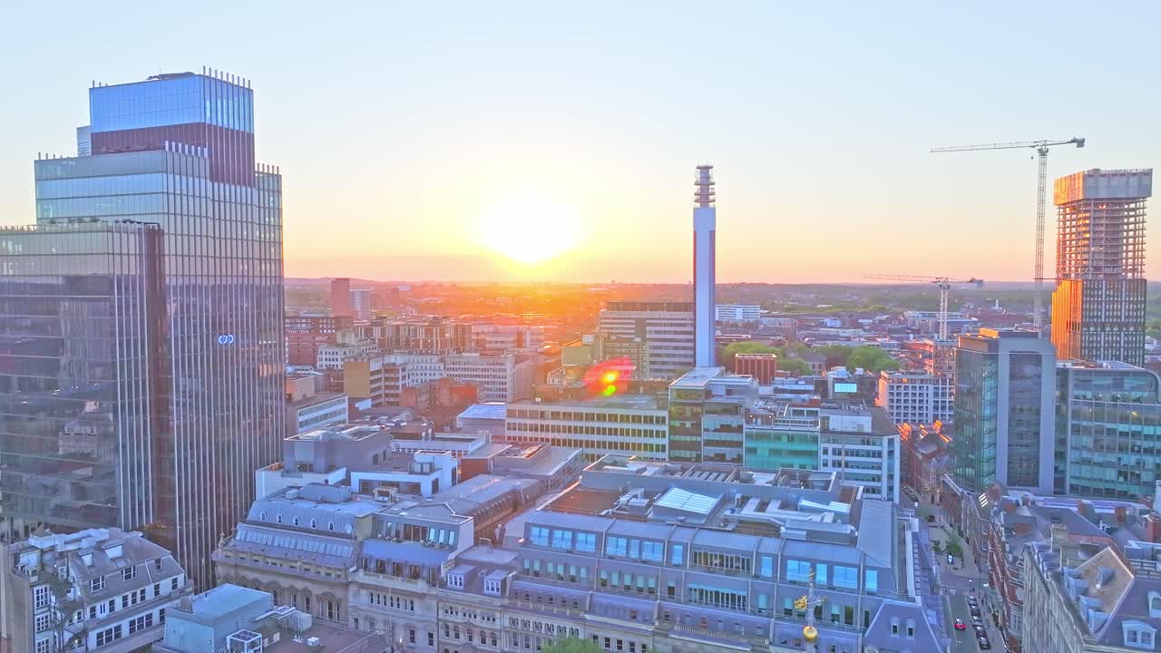 Drone footage reveals the Birmingham skyline at sunset, featuring the BT Tower, modern glass buildings, historic architecture, and cranes over a glowing cityscape