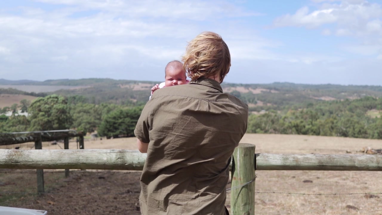 A man with long hair holding his new born baby on a farm.