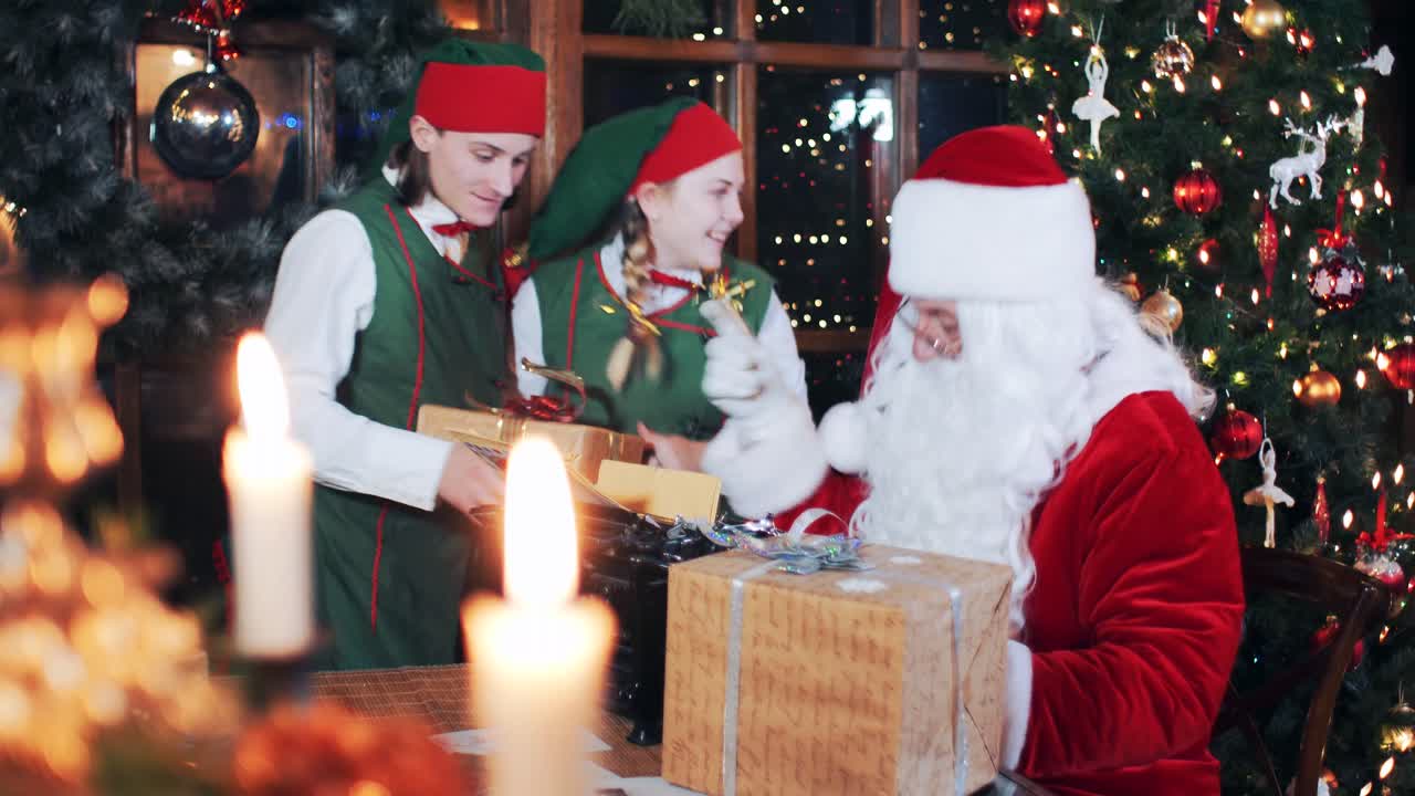 Santa Claus and elves in the evening in the room with New Year's decorations prepare parcels with gifts for children