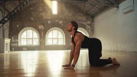 a man in a black sports summer uniform does cat stretch pose exercises in a hall that is consecrated by the rays of the sun