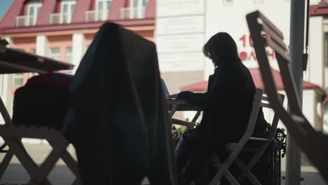 freelancer sitting outdoors at cafe table working on laptop surrounded by wooden chairs and potted plants, with a view of people walking in the background and building facades in the distance