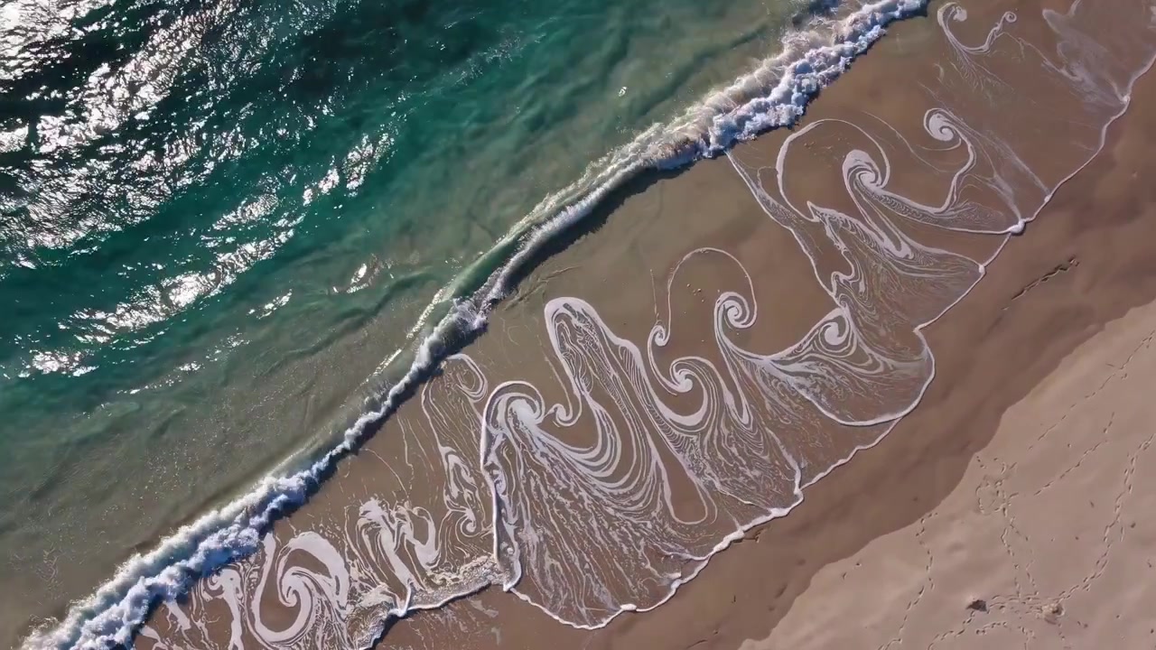 Aerial View of Ocean Waves Creating Swirling Foam Patterns on Sandy Beach