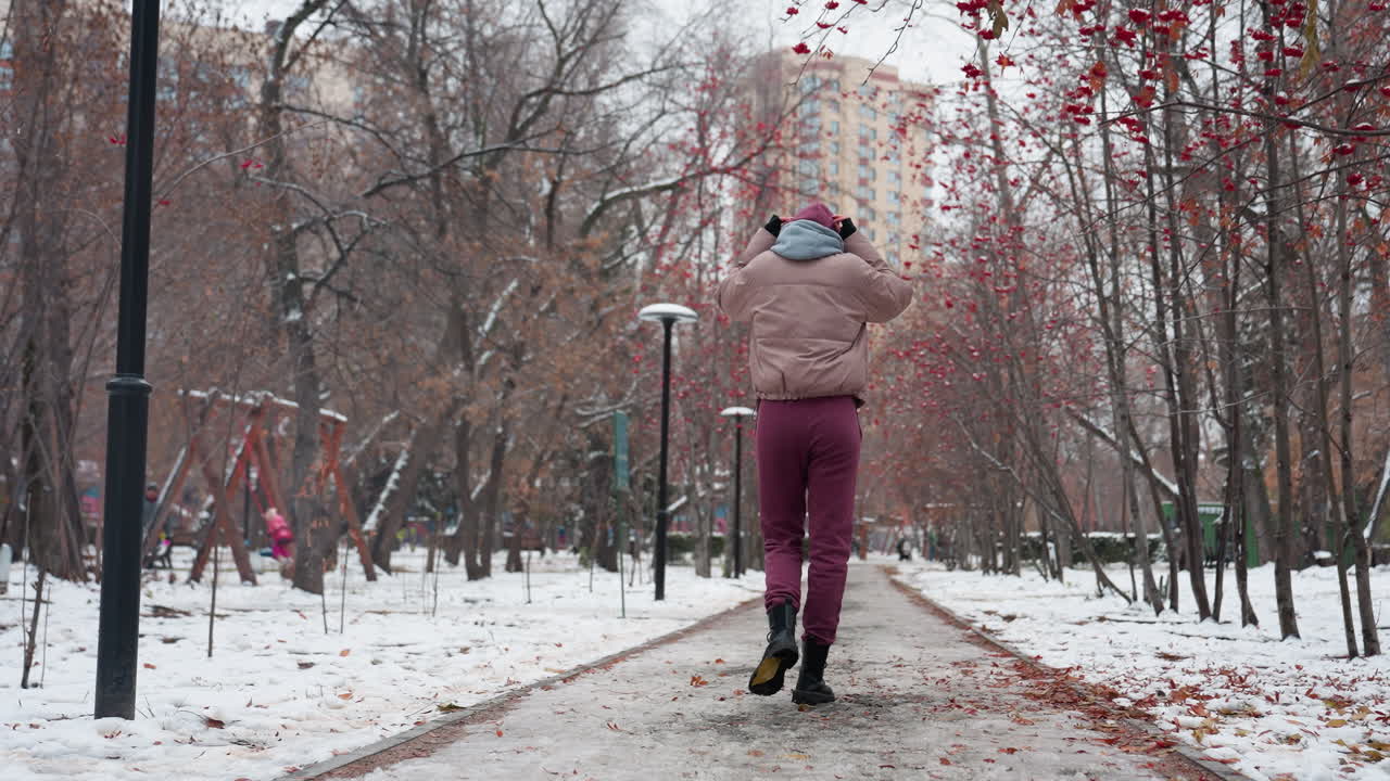 눈 인 땅, 벌거벗은 나무, 그리고 배경에 놀이터와 함께 겨울 공원을 걸어가는 여자의 뒷면, 냉정한 삶에 생명을 더하는 스에있는 아이가 보입니다.