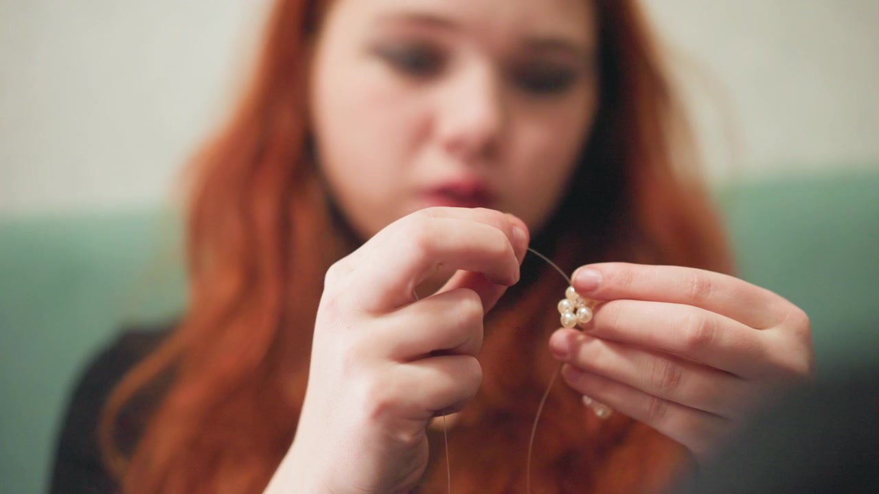 Redhaired white woman assembling necklace, hands and face in frame, concentrated expression on teal sofa, beads on string, visible scissors, DIY crafting vibe, tutorial energy and careful technique